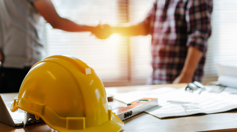 Hard hat on desk with two people shaking hands behind to support Turner Construction article