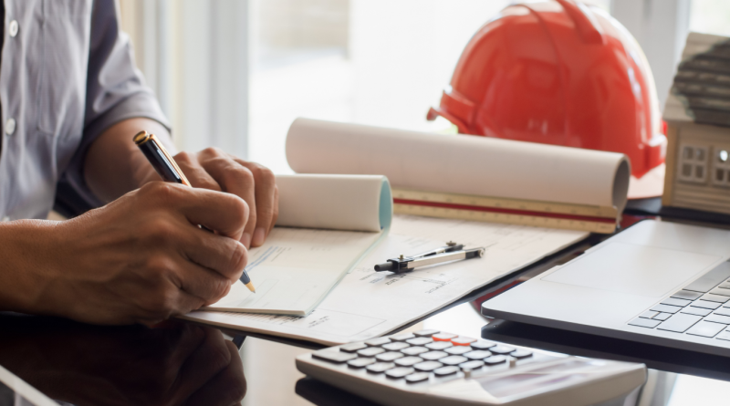 Construction payroll administrator writing a check with construction plans, calculator, and hard hat in view, illustrating construction payroll software workflow.