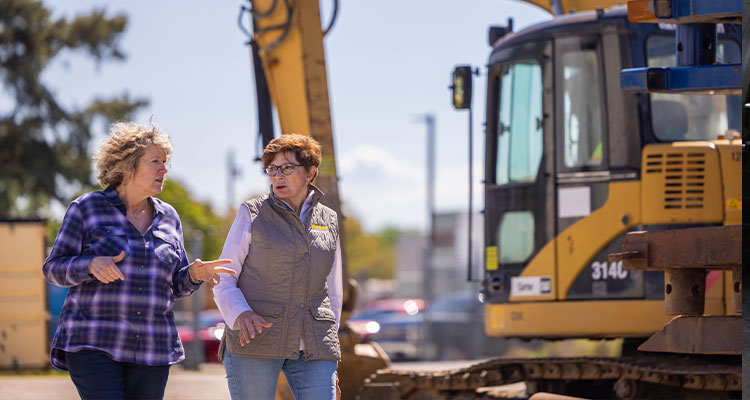 Learn how Hudgins Contracting has evolved to champion women in construction two women standing in front of a large yellow excavator