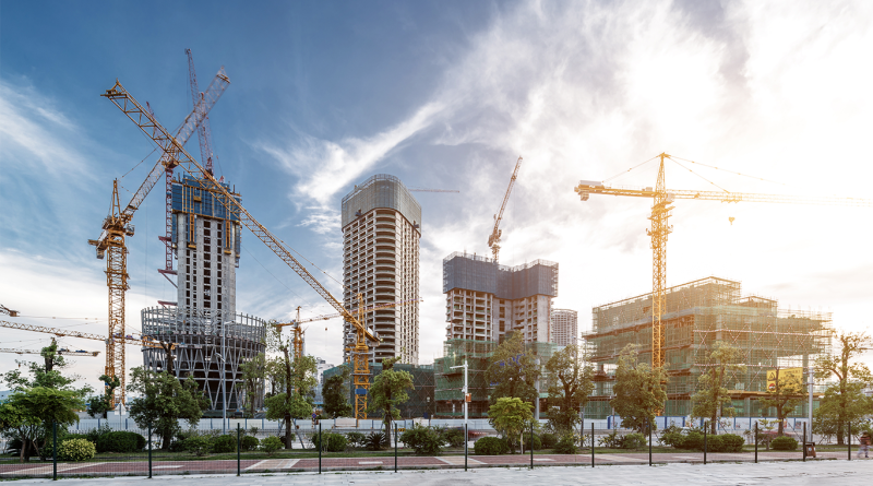 A bustling urban construction site featuring several high-rise buildings at various stages of completion, with multiple yellow tower cranes positioned against a bright, hazy blue sky.