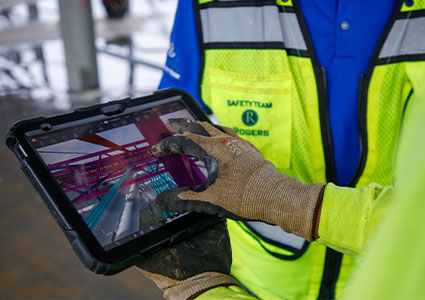 A person wearing a blue shirt, a high-visibility yellow and green safety vest, and work gloves is interacting with a rugged tablet