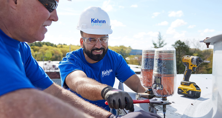 individuals working on an industrial unit while wearing safety gear, including a hard hat branded with Kelvin Group