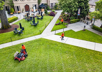 A high-angle, outdoor shot shows a crew of landscapers in bright orange safety gear maintaining a large, lush green lawn surrounding a paved courtyard