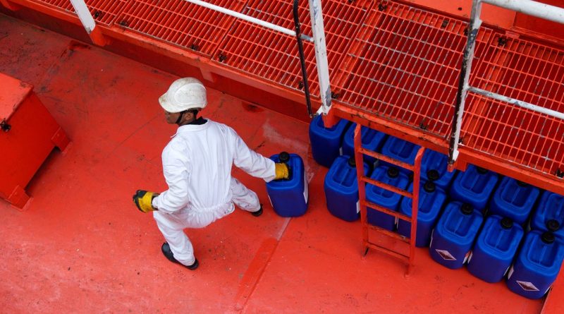 Maritime worker in protective clothing carrying a blue container across a red ship deck
