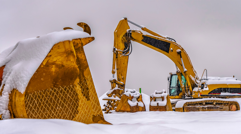 Snow-covered yellow excavator and construction equipment on a winter construction site, highlighting the challenges of working in freezing conditions.