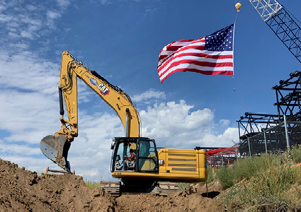 a yellow Caterpillar (CAT) hydraulic excavator working at a construction site with an American flag flying in the background