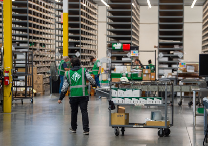 a large warehouse or fulfillment center interior with high storage racking units, a concrete floor, and workers wearing high-visibility vests pushing rolling carts or picking stock