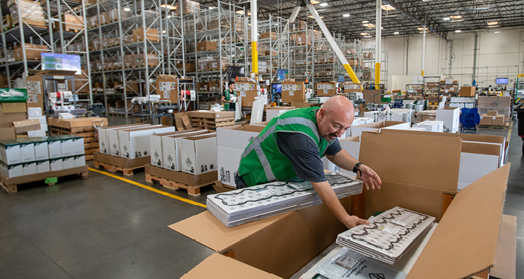 a worker in a green vest packing items into cardboard boxes inside a large industrial warehouse or distribution center