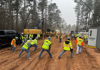 a group of construction and utility workers performing a "stretch and flex" warm-up routine on a muddy job site before starting their shift