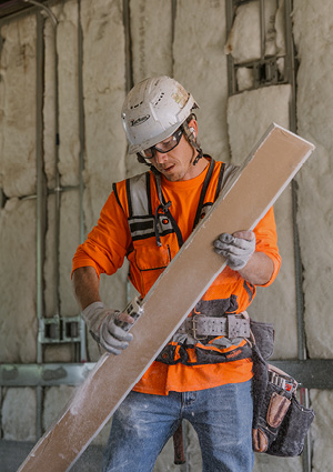 A construction worker is wearing a white hard hat, safety glasses, an orange high-visibility vest, and a tool belt while holding and working on a building material panel