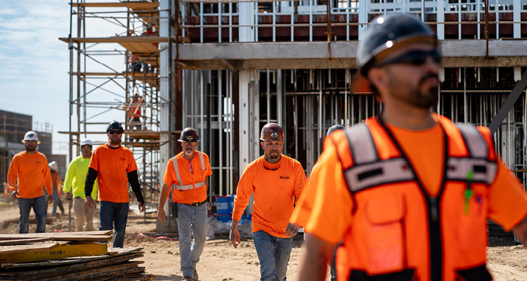 a group of construction workers in high-visibility orange shirts and safety hard hats walking at a building site with scaffolding and structural steel framing in the background
