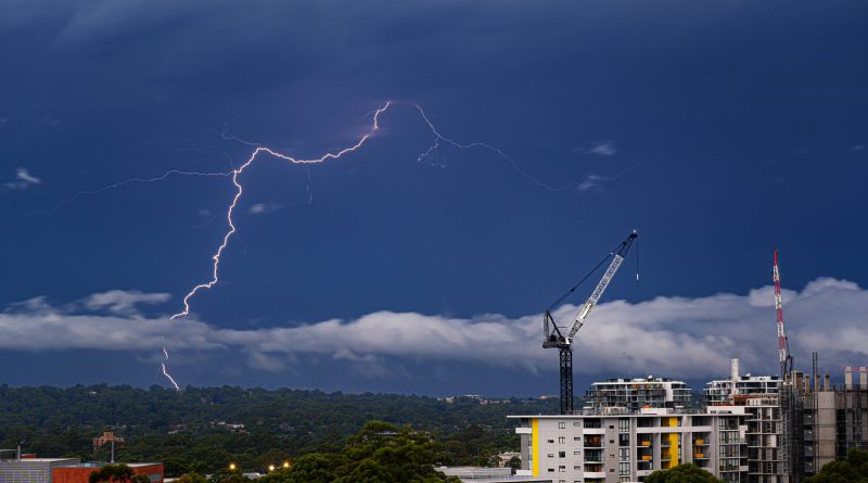 Construction cranes and residential buildings beneath a severe thunderstorm with lightning overhead.